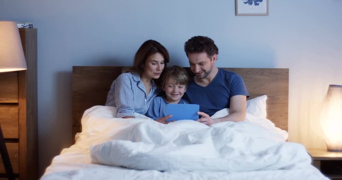 Portrait shot of the happy Caucasian parents with little son sitting in the bed before sleeping, laughing and watching something funny on the tablet device. Indoor.