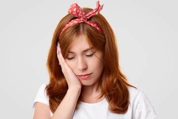 Stressful lonely displeased female closes eyes and keeps hand on cheek, feel tired of everything, being lonely and bored, suffers from not mutual love, wears headband on head and casual t shirt
