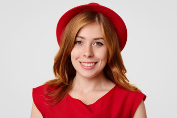 Attractive female teenager prepares to have party with friends, dressed in elegant red clothes and hat, being in high spirit, looks directly at camera with positive smile. People and happiness concept