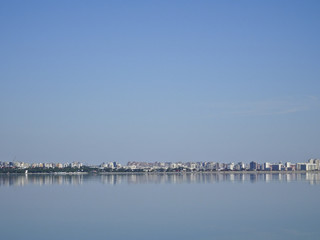 Fototapeta na wymiar Panorama of the city against a calm lake and blue cloudless sky