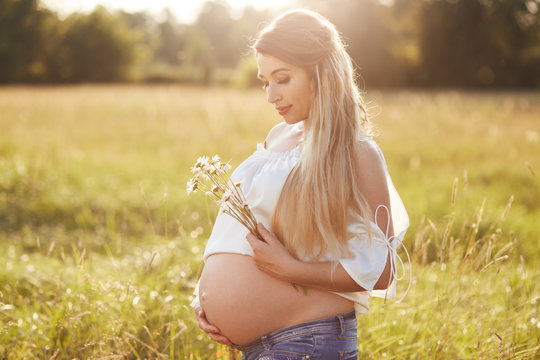 Attractive young pregnant woman shows her belly, holds flowers, enjoys sunshine and beautiful nature, stands on meadow, has long hair, waits for baby to born during summer time. Pregnancy concept
