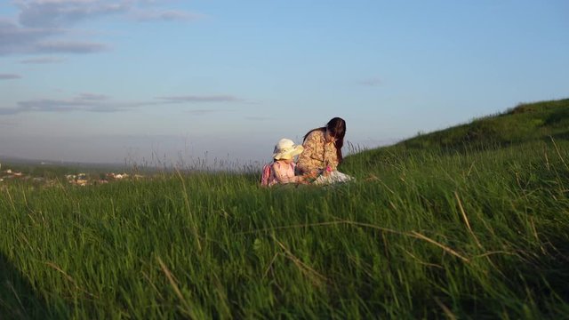 The family sits on a blanket on a picnic on a summer day on a hill with green grass. Young mother and little daughter draw.