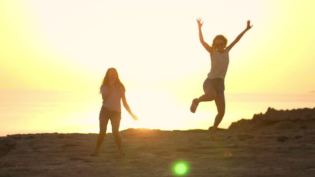 Silhouettes of woman jumping at summer sunset. Two young girls jumping at bright sunset at beach. Young girls at sunset. Cheerful human silhouettes in slow motion. Happy women enjoying evening sun