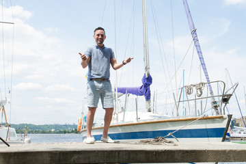 Smiling Yachtsman portrait on pier with both thumbs up. river and yachts on background. Young successful man sailor with own yacht. Man extremely have fun with yachting.