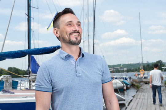 Young cheerful bearded man relaxing on sailboat posing and looking at far on background of boats, sky and folded wings. Portrait of yachtsman a fellow in blue polo sits on pier and boats on water.