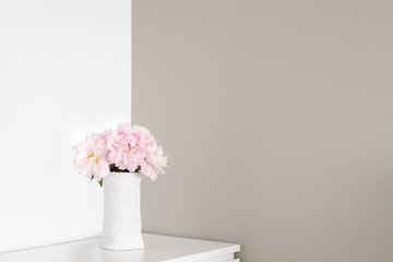 Angle of white and gray walls in room. On dresser stands white vase with pink flowers blooming...