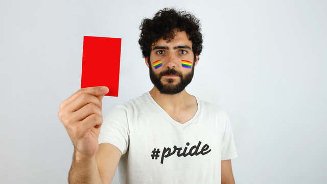 Front view of a brunette man with rainbow flag makeup in his face holding a red card. Stop homophobic concept
