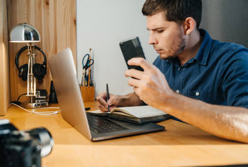 Man with a beard in a blue shirt Freelancer or photographer, with a laptop on a wooden table in his home office, writes to a notebook and talks on a videoconference.