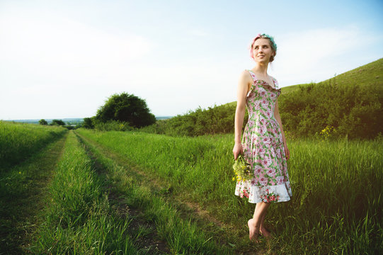 Young Happy Smiling Girl In A Calico Dress With A Bouquet Is Walking Along A Country Road With Green Grass