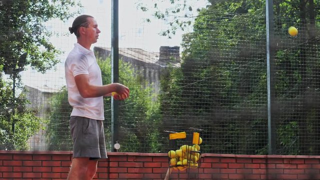 Young attractive coacher serve a tennis ball from metal basket during training