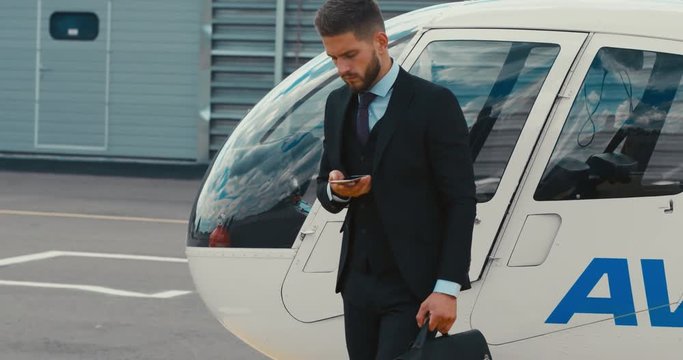 Young adult businessman executive talking on the phone near a private charter helicopter on a helipad. 4K UHD