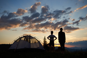 Fototapeta na wymiar Back view silhouettes of a female and a kid stand near a camping at daybreak. People enjoy the scenery of the morning mountains and hills above which the sun rises, under a fairy sky with low clouds.