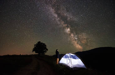 Fototapeta na wymiar Active female hiker resting at night camping in mountains under amazing night sky full of stars and Milky way. Girl standing beside illuminated tent and looking at sky full of stars. Astrophotography