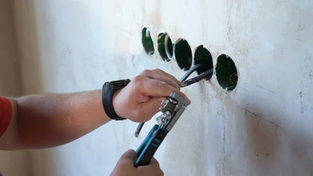 Male worker with pliers cleaning wires in junction box for socket. Closeup hands preparing cable for equipment installation on building site. Safety policy, technology, repair, interior design concept