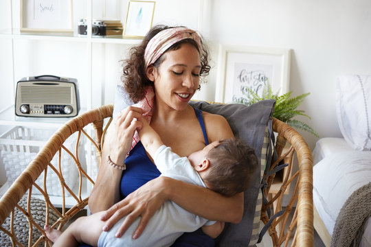 Sweet shot of happy young woman of mixed race appearance sitting in chair, breastfeeding her infant son, enjoying deep connection with child. Love, family, relationships and togetherness concept