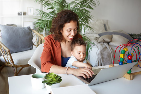Indoor shot of cheerful young mixed race self employed woman using portable computer for remote work, has to babysit her infant son and check email simultaneously. Maternity and freelance concept
