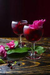 Two glasses full of red drink with berries, ice and a pink flowers, beside which lie berries and a branch of green tropical leaves on a vintage wooden table