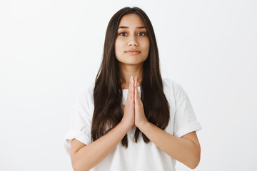 Studio shot of grinning good-looking female coworker in white t-shirt, holding hands in pray and smiling with pleased expression, asking for offer or begging for help, apologizing over gray wall