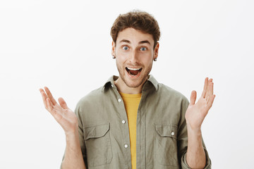 Fototapeta premium Amused good-looking happy male with beard and short curly hair, clasping in hands liking perfect perfomance of favorite band, smiling broadly and gazing with delight at camera, posing over gray wall
