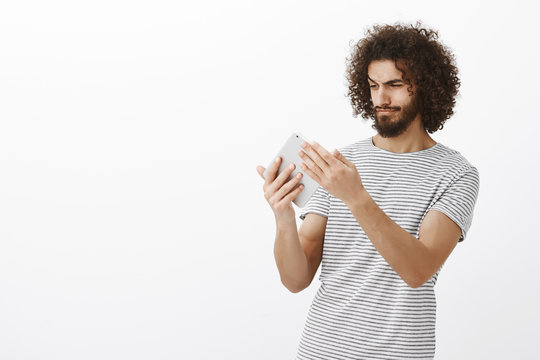 Portrait Of Creative Hispanic Male Designer With Stylish Haircut And Beard, Holding Digital Tablet And Looking Focused At Screen, Drawing Plan Of Apartment To Create Design, Standing Over Gray Wall