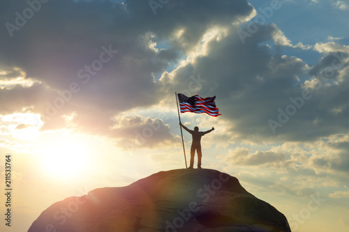the man is standing on the top of the mountain, holding his hands up with the flag of the US america against the sunset. Business concept idea, success and achievements, career ladder, victory.
