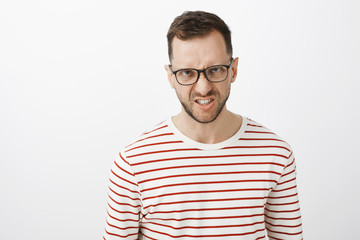 Obraz premium Hate you with all heart. Portrait of outraged disdaining european guy in black glasses, looking from under forehead and frowning, showing teeth in anger, standing over gray background