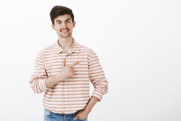 Sharing nice coffee spot with friends. Studio shot of friendly joyful caucasian guy with moustache, holding hand in pocket and pointing right with forefinger, smiling broadly, showing direction