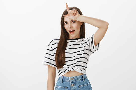 Losers and outsiders should stay home. Portrait of popular attractive female student in striped t-shirt holding l word over forehead, mocking or laughing over rival who lost bet over white background