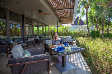 Young woman looking at the menu sitting in outdoor restaurant on background of palms and greens