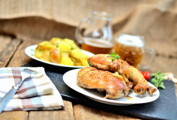 Golden grilled chicken legs on a plate, roasted potatoes and jug of cold beer, rosemary, tomatoes, oregano, salt, pepper and garlic on a wooden table - limited DOF