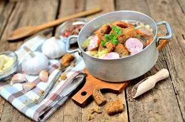 Garlic soup in an old saucepan, fresh garlic bulbs, cloves, colored pepper, wooden spoon, croutons and a towel on a rustic wooden table