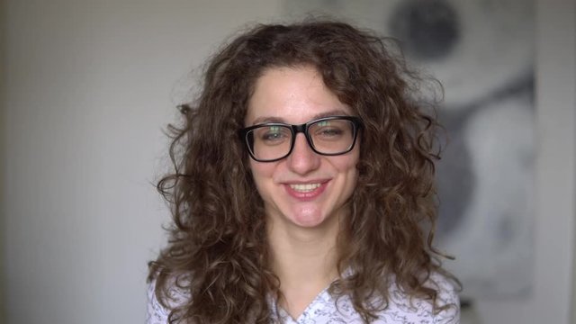 curly-haired girl wears black glasses, smiles and laughs.close up. against the white room. Skye's wearing a white shirt.