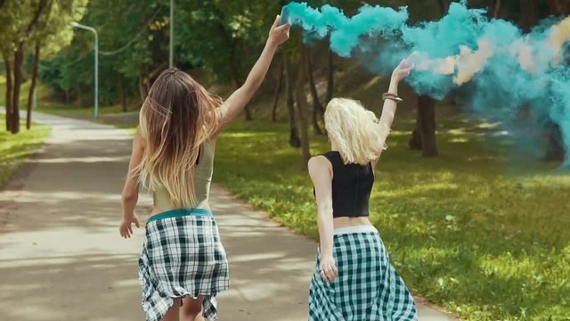 Happy hipster girls in beautiful clothes runs in park waving colored smoke. Young girls have fun and jump outdoors in slow motion. Party concept