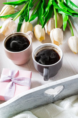 Two cups of coffee on a wooden tray with flowers and a gift box on a bed with a blanket. St. Valentine's Day