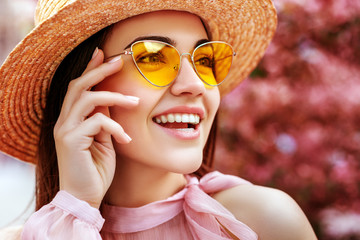 Outdoor close up portrait of young beautiful happy smiling woman wearing yellow sunglasses, straw hat, posing in street. Copy, empty space for text