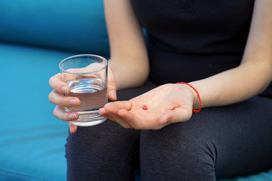 Sick woman holding pill glass of water on the sofa. Depressed unhealthy woman, about to take antidepressant pill, emergency contraceptive, painkiller for painful periods. The red thread of Jerusalem.