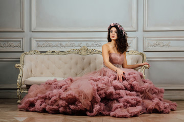 Luxurious sexy girl in an evening cloudy dress sitting on a sofa in the studio interior.