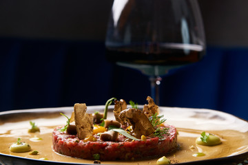 Beef tartare Steak with egg yolk, pistachios, truffle oil, green mayonnaise on porcelain plate and glass of red wine on restaurant table. Classic tartare meat with ingredients. Luxury food