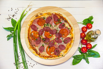 appetizing pizza with sausages and ham, olive oil and basil on a white table, flat lay