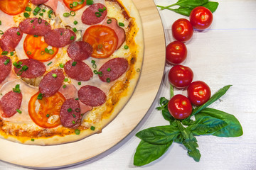 appetizing pizza with sausages and ham, spices, basil and green onions on a white table, flat lay