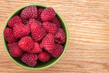 Top view of fresh red raspberries in a brown bowl on a light wood background
