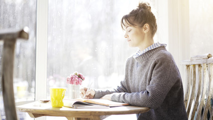 Obraz na płótnie Canvas A profile of a smiling young brunette girl in a cafe sitting near the window dressed in a blue pullover is reading a notebook in the cafe writing with a pencil.