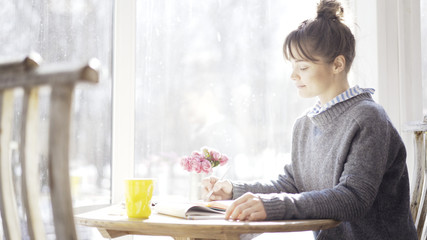 Fototapeta na wymiar A smiling calm young brunette girl in a cafe sitting near the window dressed in a blue pullover reading a notebook holding a pencil