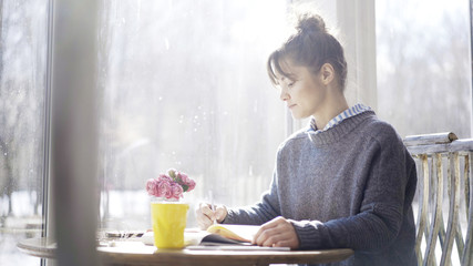 Obraz na płótnie Canvas A young serious brunette girl dressed in a blue pullover is writing down notes to a notebook in the cafe sitting near the window