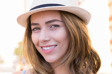 Close up portrait of blonde young woman with white hat looking at camera outdoors in the streets of the city. Bright day light. Concept of happiness, style, confidence.