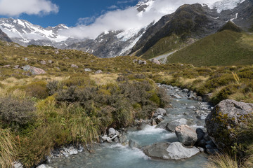 Naklejka na ściany i meble Small, babbling mountain stream in Aoraki/Mount Cook National Park, seen from the Hooker Valley Track. Snow capped mountains in the background.