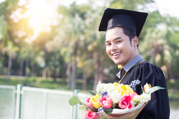 Attractive Asian graduated man in cap and gown smile and holding bouquet feeling so proud and...