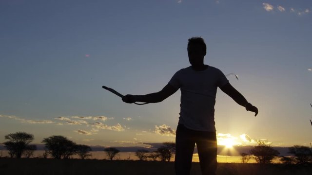 A medium shot of a silhouetted man whipping his whip out on a field with trees in slow motion.