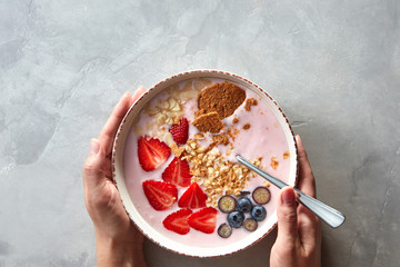 The girl's hands hold a plate of yoghurt, granola and berries on a gray concrete background. Top view