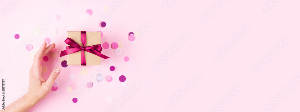 Wall mural woman hand and kraft gift box with pink bow on pink background decorated with confetti.. top view, h